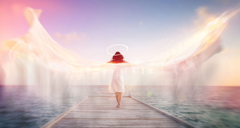 Sconceptual image of a female angel standing barefoot on an ocean jetty in a white dress with a halo and outspread wings showing motion blur with ethereal colorful sun flare effects